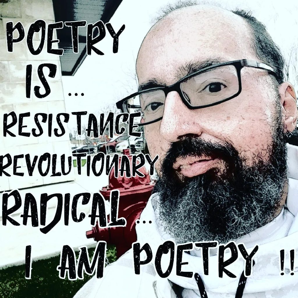 A selfie of Val, a power wheelchair user, with text that reads "Poetry is... Resistance Revolutionary Radical... I am poetry!!". He is a lightly tanned skin Latinx male appearing with short dark hair and salt and pepper beard. They are wearing black framed tinted glasses.