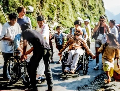 A photo shows Gene in his late-20s on a South American expedition with other individuals with disabilities. He sits in a manual wheelchair and wears 80s-style attire, large sunglasses, and a shade hat with his arms held loosely to his sides. His care attendant pushes him up a rough paved road.
