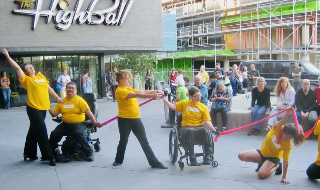 Group of people wearing yellow shirts and stretching a rubber band between them. The group has people with and without disabilities.