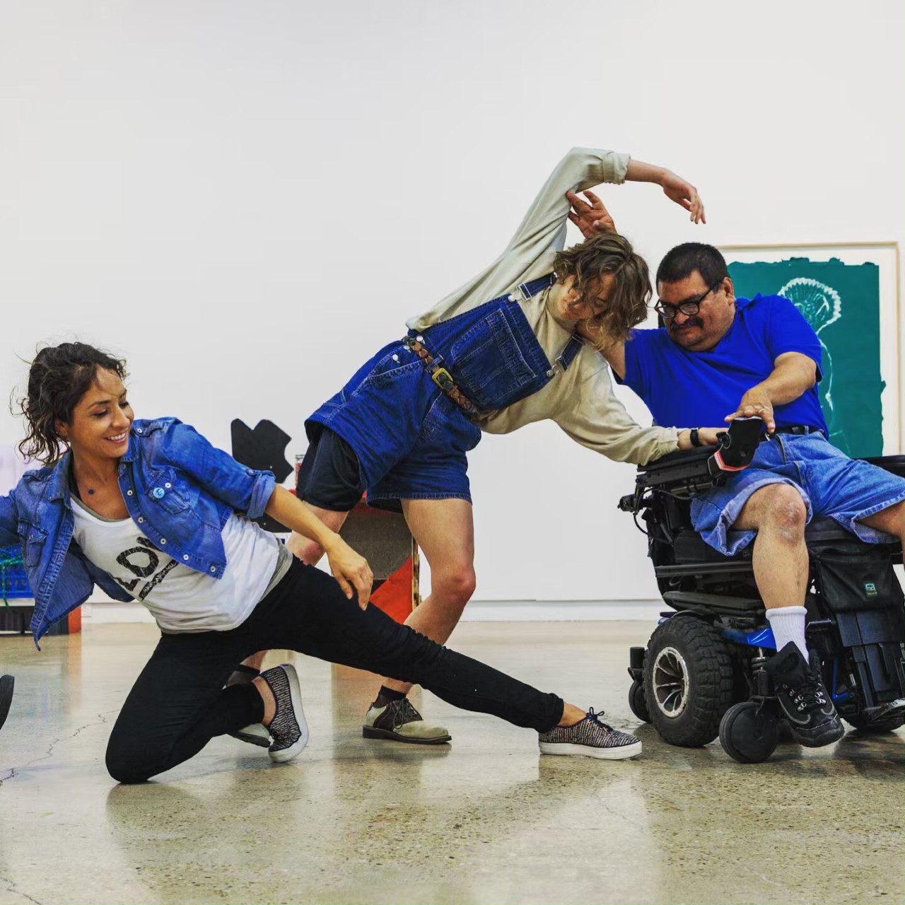 Photo taken in an indoor gallery space shows a man in a power wheelchair dancing with a woman arching her back and arms towards him. A woman in the foreground extends her leg between the two.