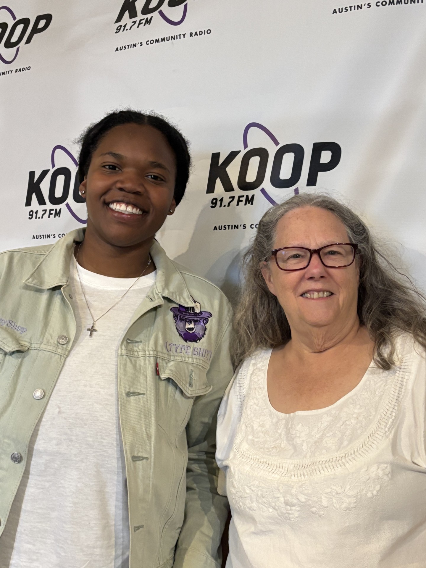 Jaela Mitchell, Art Spark Texas' radio hour intern smiles with Executive Director and radio show host Celia Hughes in front of KOOP radio's backdrop in the studio