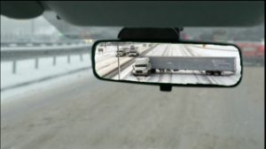 A tractor-trailer jack-knives on a snowy freeway, as seen from a rearview mirror