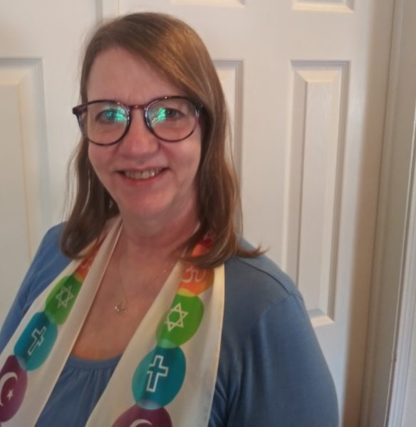 A photo of Kaye with light brown hair smiles into the camera. She is wearing a multi-colored stole with interfaith symbols and a blue shirt and dark-rimmed glasses.