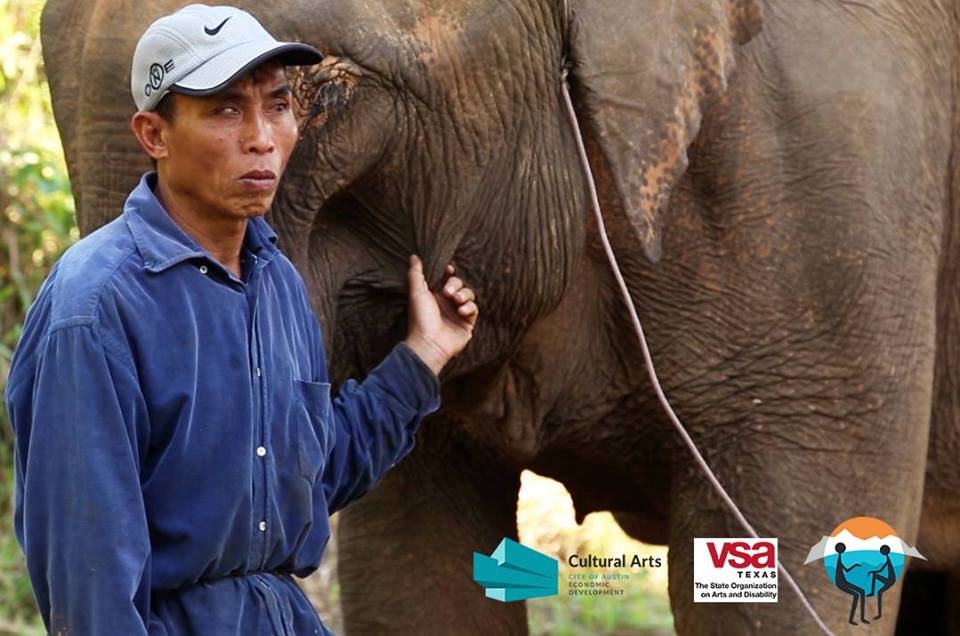 Older Asian man in dirty blue work clothes gently pinching the cheek of a large elephant