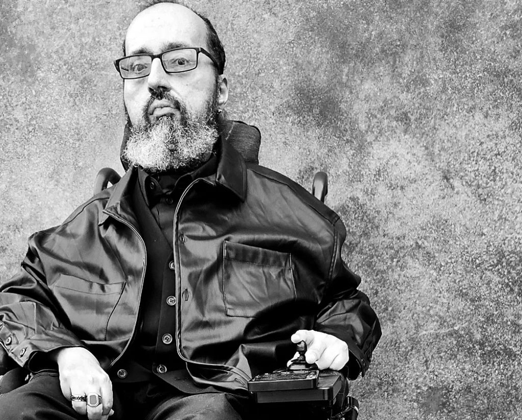 Black and white portrait of Val, a powerchair user, with one hand on the joystick, parked in front of a textured wall, looking slightly down at the camera. He is a lightly tanned skin Latinx male appearing with short dark hair and salt and pepper beard. They are wearing all black and have black glasses.