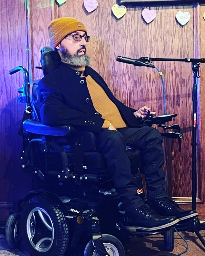 Val, a powerchair user, speaking into a microphone, with Valentine's day hearts strung across the wall behind him. He is a lightly tanned skin Latinx male appearing with a salt and pepper beard. They are wearing an orange beanie and shirt and black pants and jacket.