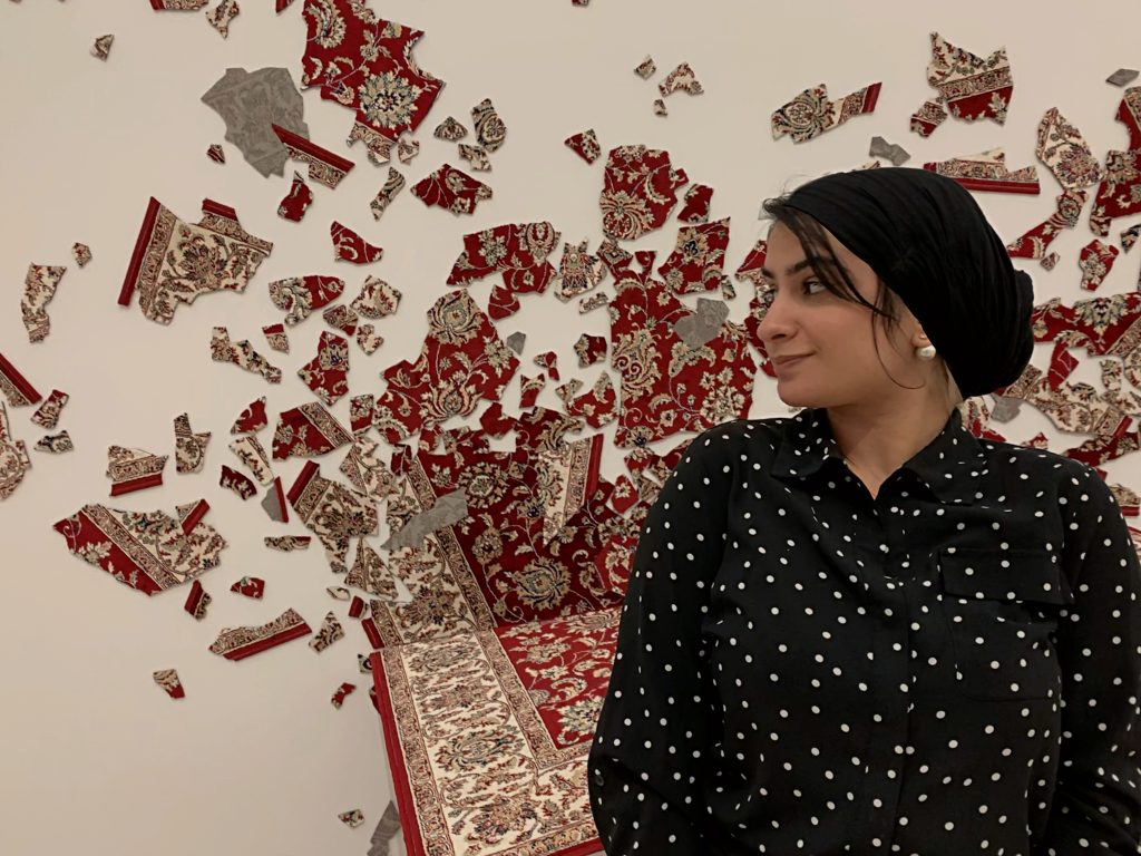 Rouqayya posing beside a work of art in which a carpet appears to be shattering against a wall.