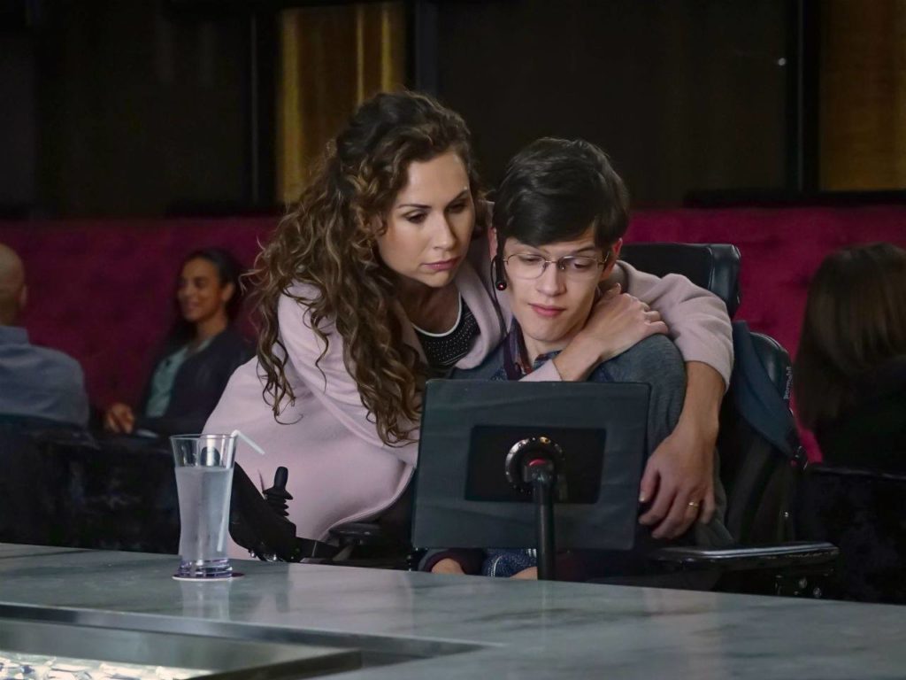 Still image from TV show Speechless, in a restaurant, mother leans protectively over teenage son in a power wheelchair.