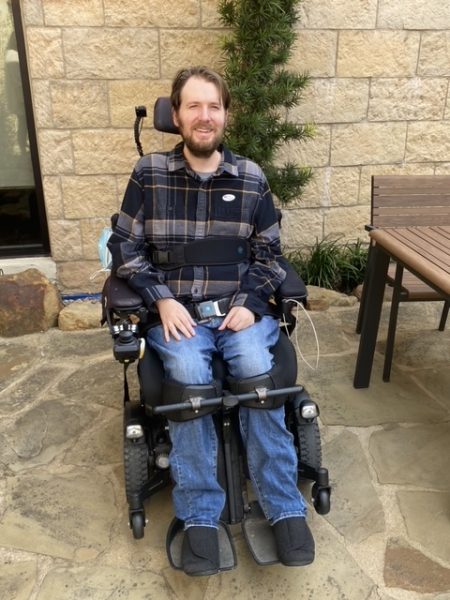 Eric smiles while posing for a photo in his power wheelchair. He wears an “I voted!” sticker on his shirt.