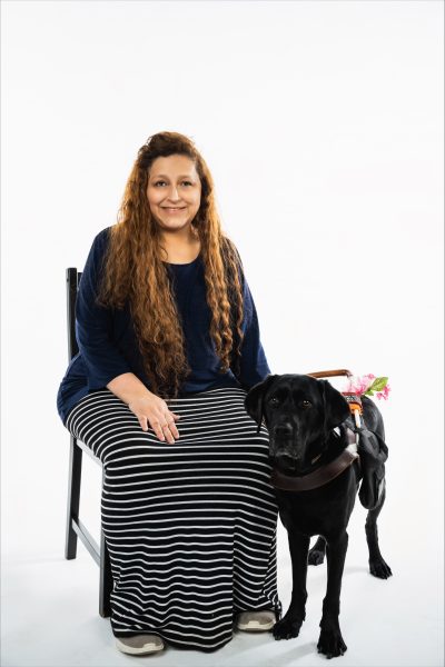 Yadira smiles into camera. She has long, wavy brown hair and wears a black and white striped skirt and black long-sleeved top. She holds Deja’s harness who stands on Yadira’s left side touching her knee. Deja looks into the camera. 