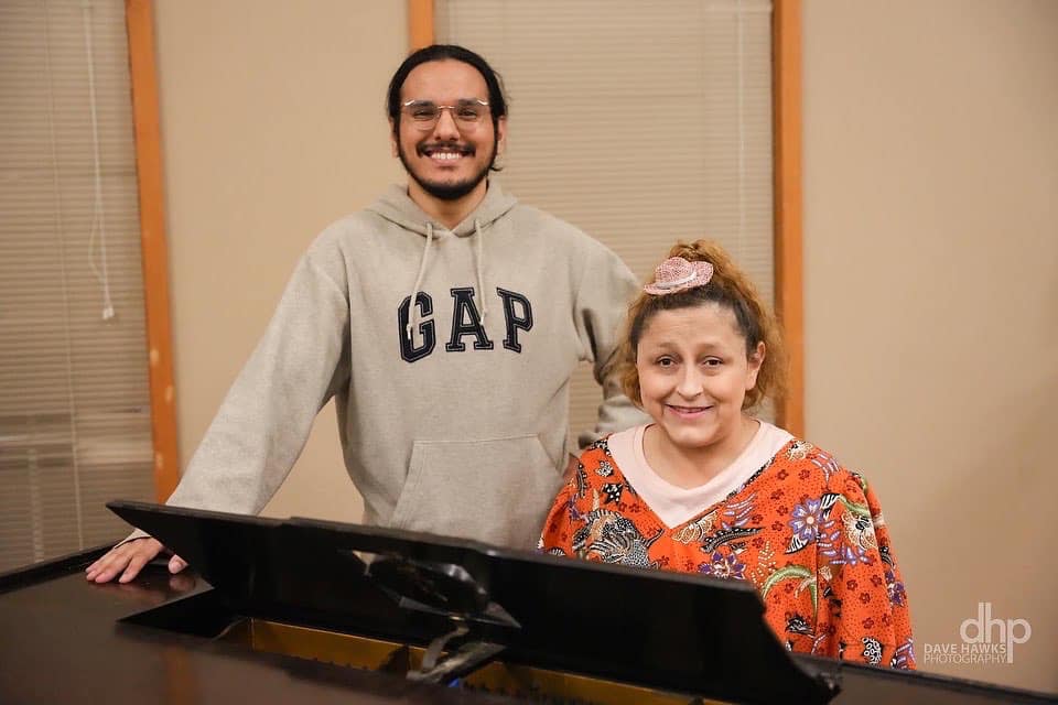 Yadira sits at grand piano smiling into the camera. She has curly brown hair tied up behind her head with a bright orange shirt with an exotic bird pattern. Micheal Galvan, her brother in Strange Faces, stands behind her slightly to her left.