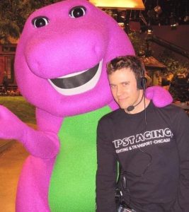 Person standing next to Barney the Dinosaur, a purple dinosaur mascot costume. 