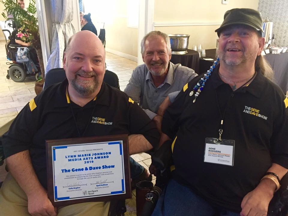 A photo of Gene and Dave posing with their freshly-awarded Lynn Marie Johnson Media Arts Award at the 2019 Art Spark Artist of the Year Awards. They both smile while seated in their power wheelchairs and wearing official “The Gene and Dave Show” golf shirts. Their friend, Coalition of Texans with Disabilities Executive Director Dennis Borrel, smiles while posing between the two.