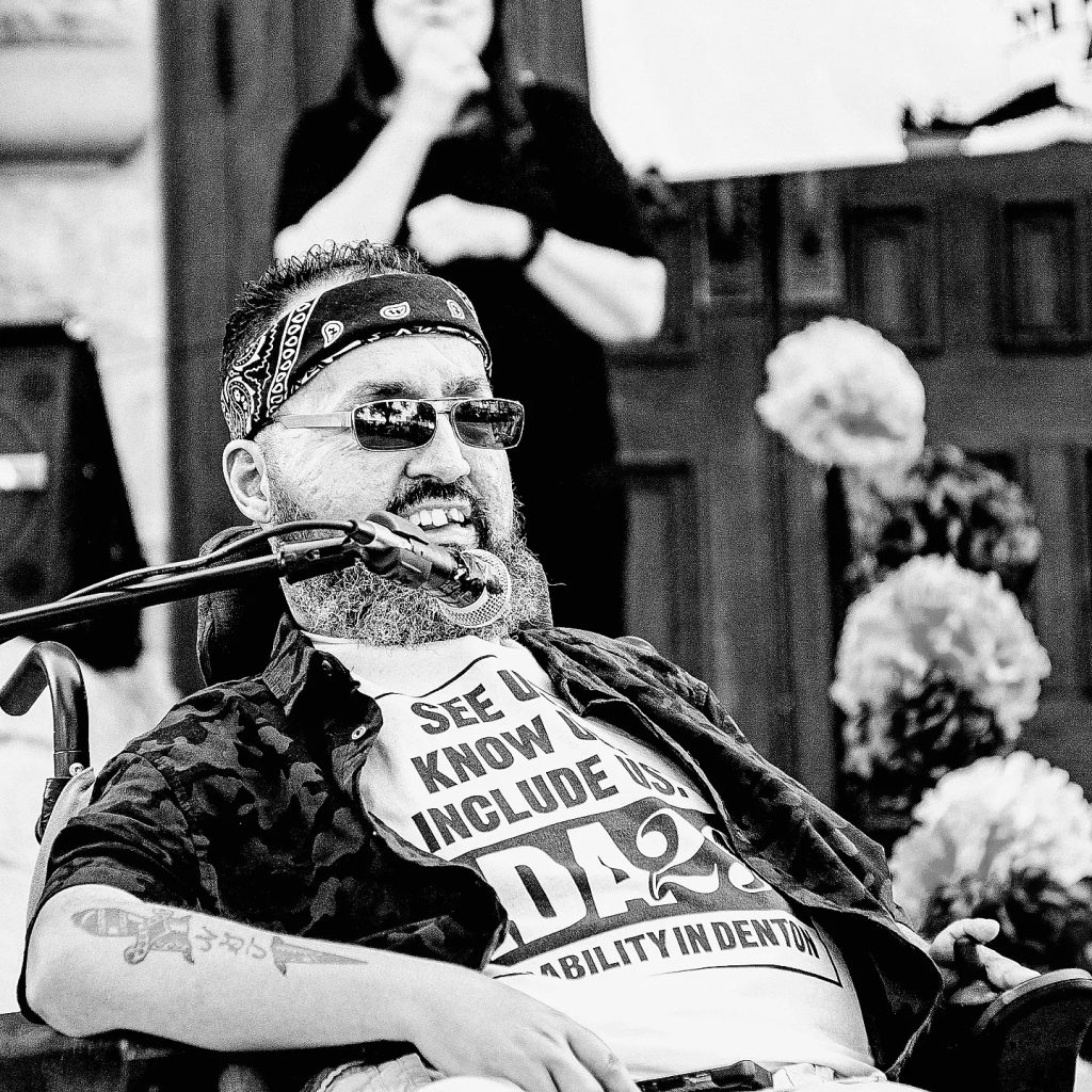 Black and white photo of Val, a powerchair user, speaking into a microphone, with flowers and an ASL interpreter blurred out in the background. He is a lightly tanned skin Latinx male appearing with short dark hair and salt and pepper beard. They are wearing a dark bandana on their head, dark sunglasses, and white ADA28 t-shirt under a dark jacket. The sleeves are rolled up to reveal a knife tattoo on one arm.