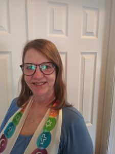 A woman with circular framed glasses and shoulder length brown hair smiles for the camera. She is wearing a scarf with a variety of religious symbols on it.