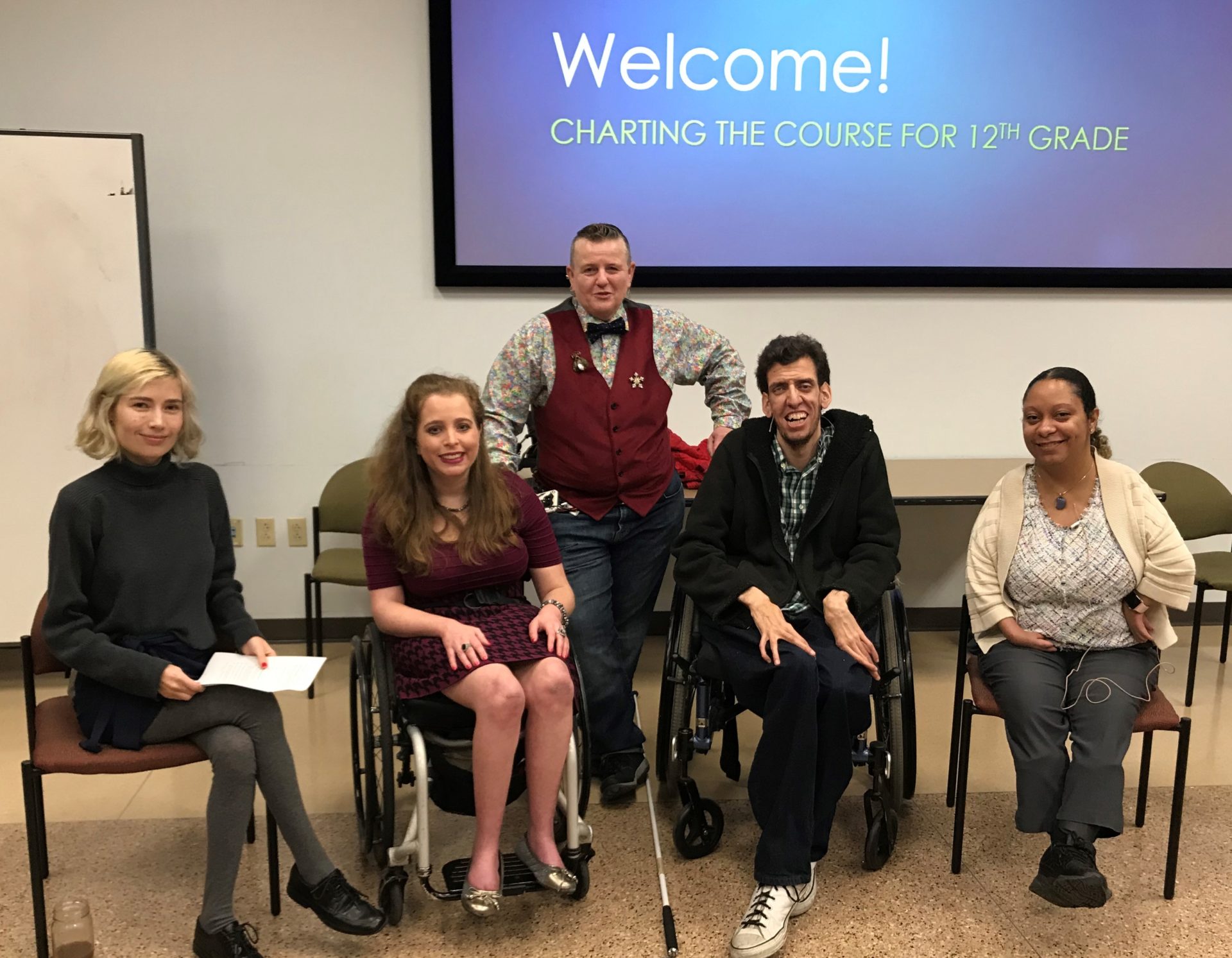 A photo of 5 adults with various disabilities smile in front of a projection screen.