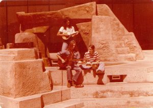 Sepia toned photo of three musicians busking on the streets of 1970s Denver. In the front, a guitar player and a bongo drum player sit side by side. In the background, a young Joanne stands, playing mandolin.