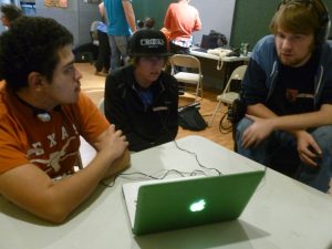Photo of a volunteer instructing two young adults who are seated at a laptop in a large classroom.