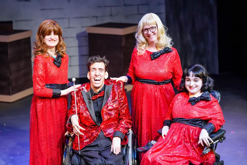 A photo of Kaye on stage with the cast of Pandora, a TILT Performance production. Kaye is wearing a long blond wig and a bright red sequined full gown.