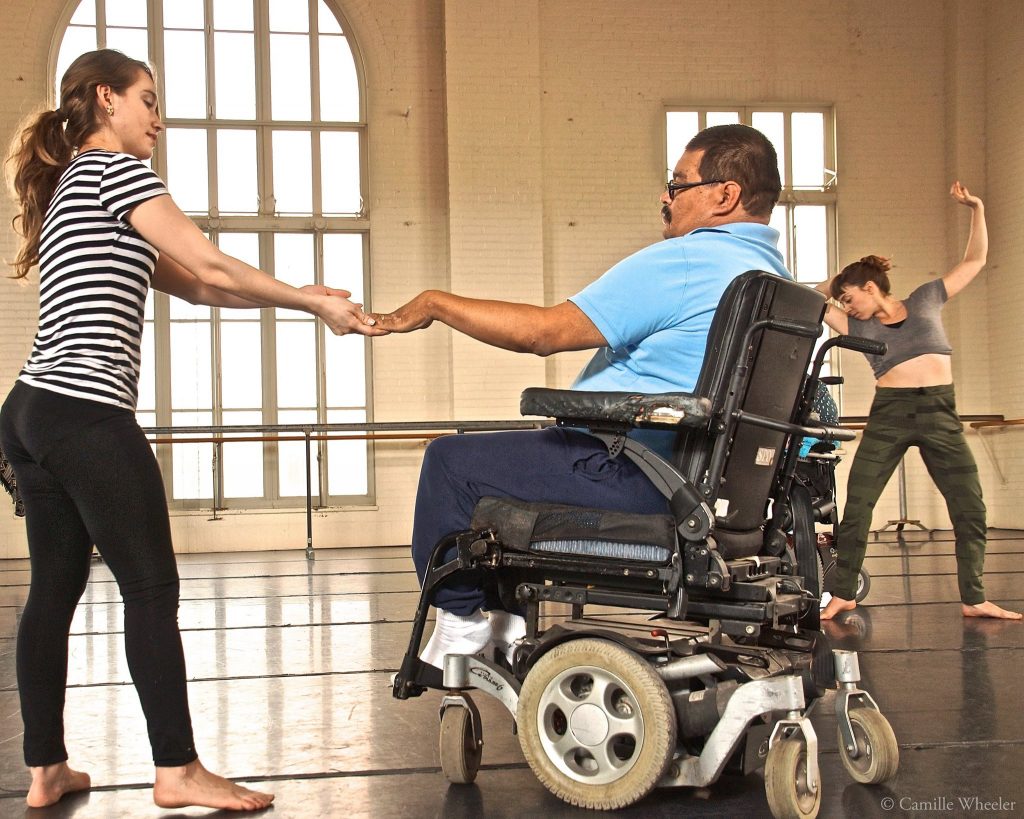 a man in a wheelchair holds hands with an able-bodied female dancer in a dance studio