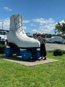 Zoe is leaning on the world’s largest roller skate. They are wearing dark sunglasses and their arm is bent to rest their hand on their face.