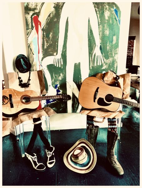 Two flattened paper bags are propped up in chairs. They are accessorized with shoes, hats, and jewelry and have guitars in their laps