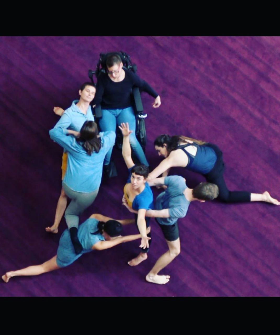 Photo taken from above where people with and without disabilities have created a sculpture. Everyones gaze is up towards the camera. All the dancers are wearing clothes in different shades of blue and black.