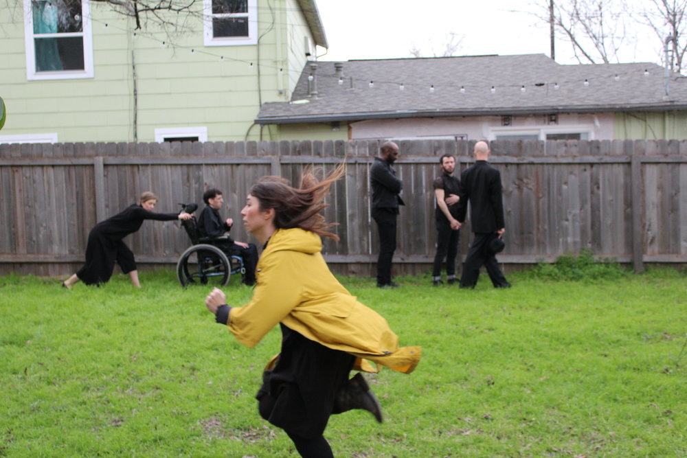 Antigonick cast dressed in black and lined up in front of a fence