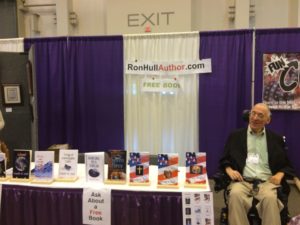 author Ron Hull sits in his power wheelchair beside his booth of books and a sign hanging behind him reads ronhullauthor.com