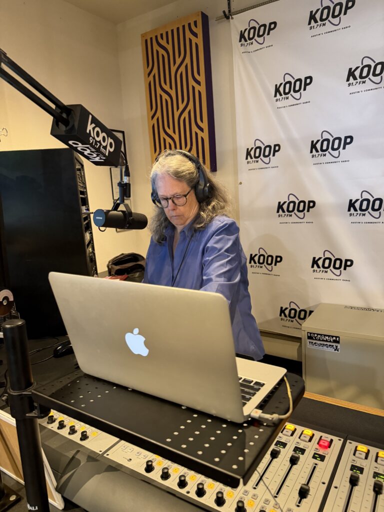 Celia Hughes, Director of Art Spark Texas, starts her show on KOOP in the studio standing at the mic infront of a black and white Koop radio backdrop