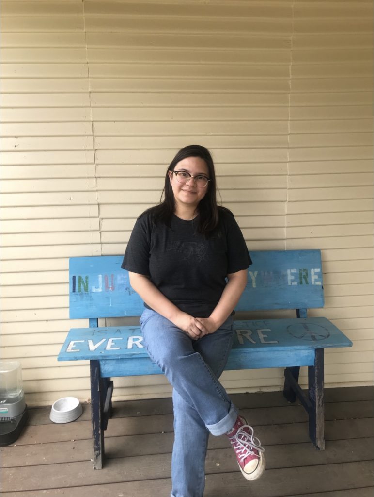 A photo of Diana smiling at the camera while seated on a blue painted bench on the Art Spark office porch