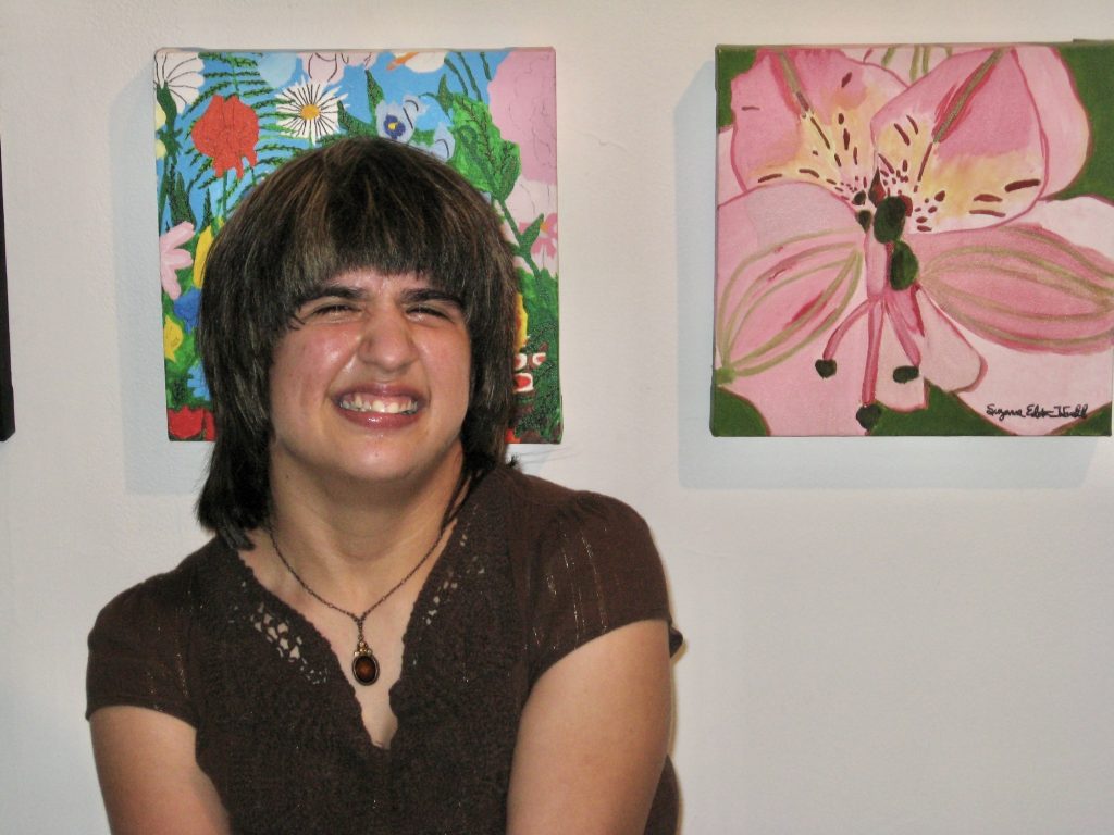 An artist smiles in front of a wall where their artwork is hung.