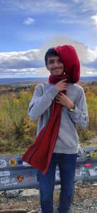A young man with short black hair and mustache poses on the side of the road overlooking a field of wildflowers. He is posed facing the camera smiling while holding his long red scarf that is blowing in the wind. 