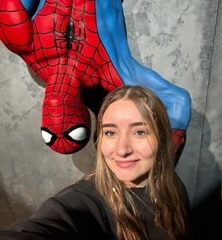 A headshot of Megan Reid standing in front of a Spiderman sculpture that is hanging from the ceiling.