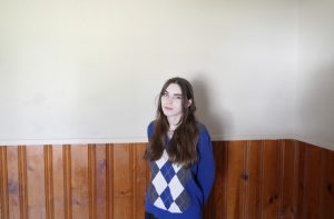 A girl with long brown hair poses with her arms behind her back in front of a wood paneled wall. 