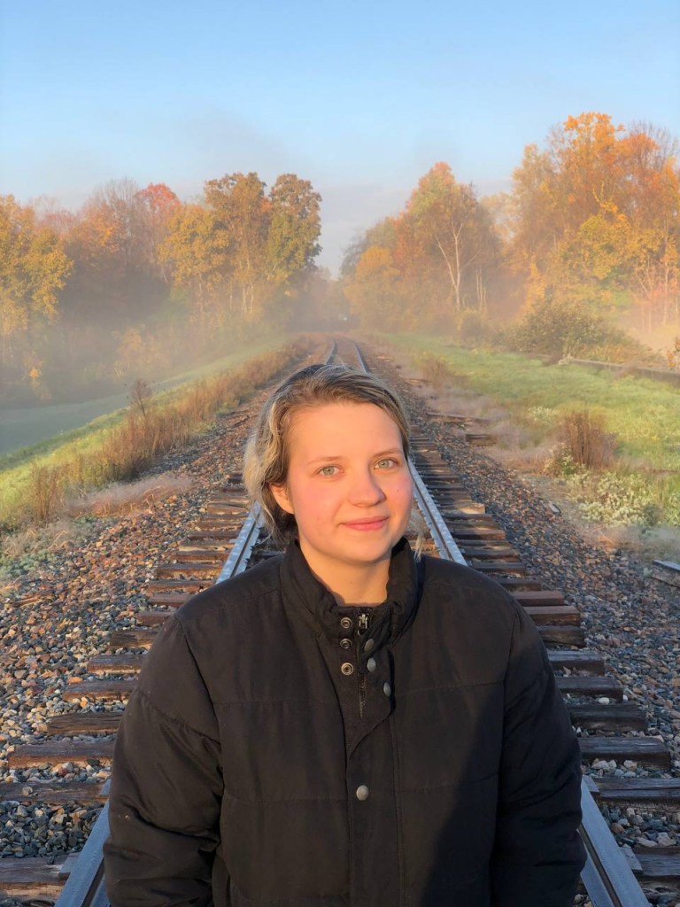Veronika Kostyaeva stands on railroad tracks in front of a foggy forest. 