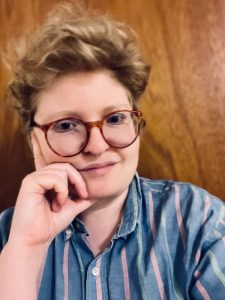 Photo of Joey Gidseg, a white nonbinary person with short blonde curly hair and big red glasses, resting their chin on their hand with a slight smile. Joey is wearing a light blue shirt with pink and white stripes and is in front of a wood paneled background.