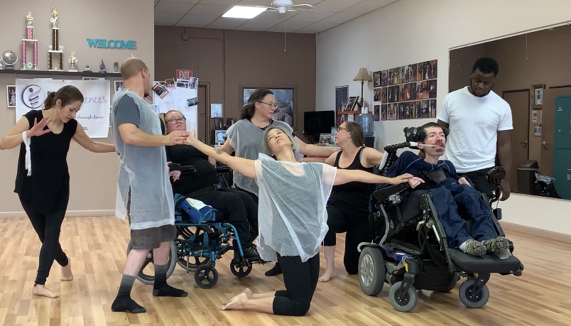 dancers with and without disabilities are making a multilevel group shape while wearing black outfit that has a see-through cape on top of it