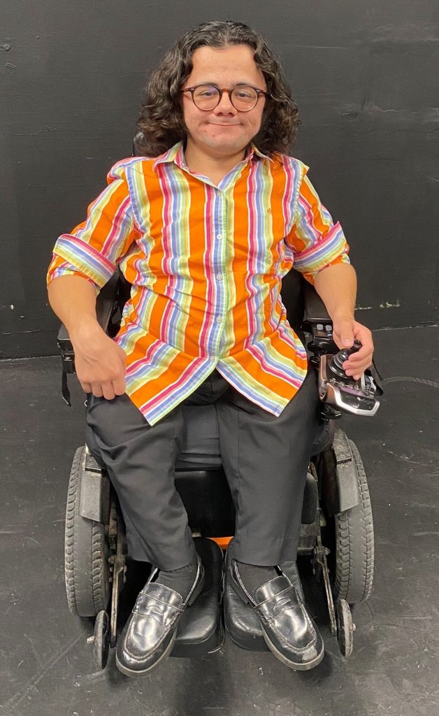 ID: Kurt sitting in electric wheelchair grins directly into camera. Wearing a bright multi-colored, striped button-up shirt and black pants. Shoulder length wavy black hair, with center parting and retro round glasses.