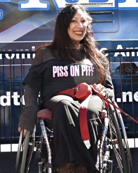 Maria poses, smiling, for a photo in front of a bus. Seated in her manual wheelchair, she has flowing black hair and wears a black dress with pink text that reads, “Piss on Pity.”)