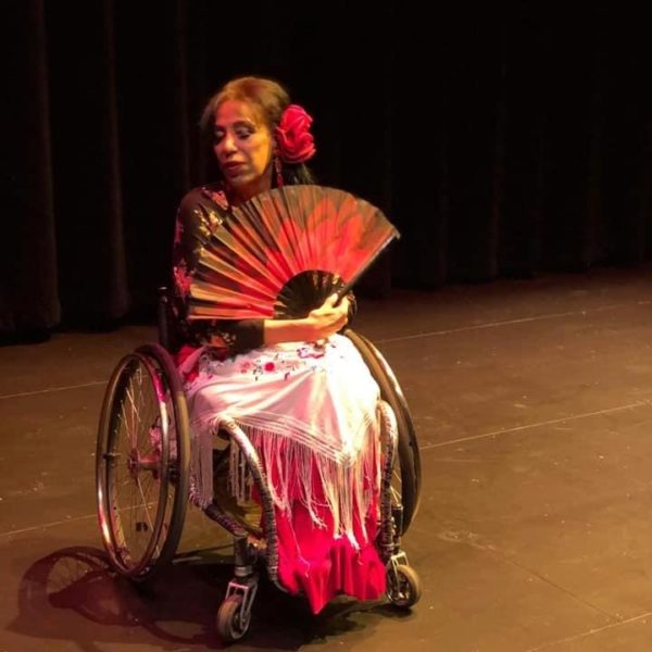 A photo shows Maria seated in her manual wheelchair on an empty theater stage, illuminated by pink lighting. She wears a red skirt and black top with a white shawl draped over her lap and a large red flower pinned in her hair. She holds a large black fan with a dark red floral design spread in front of her chest with her head turned to the right and eyes closed.)