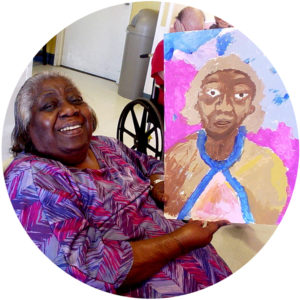 Older woman seated in wheelchair smiles as she shows off her self-portrait painting.