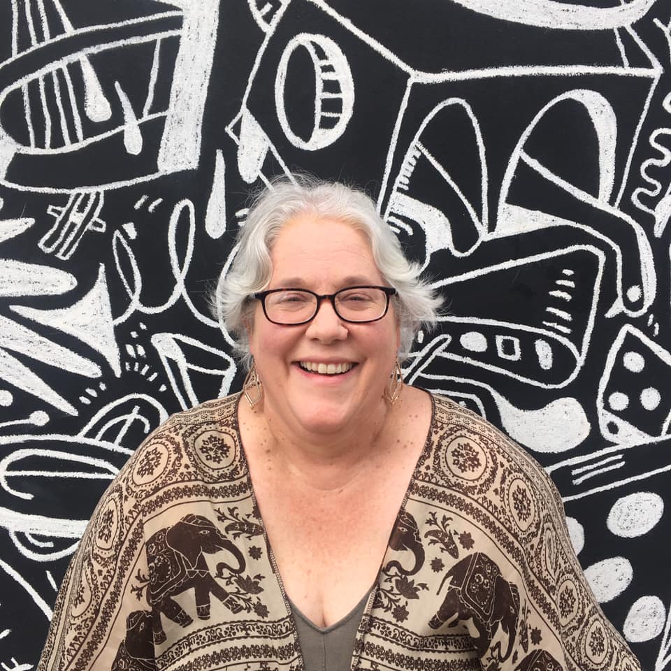 Photo of Jennifer Howell, a white woman with short silver hair. Jennifer is smiling, and wearing a beige shirt with an elephant print and black, square-frame glasses. She is standing in front of a black and white mural.
