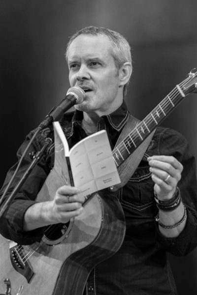 A man reads from his book of poetry at a microphone with an acoustic guitar slung around his neck.