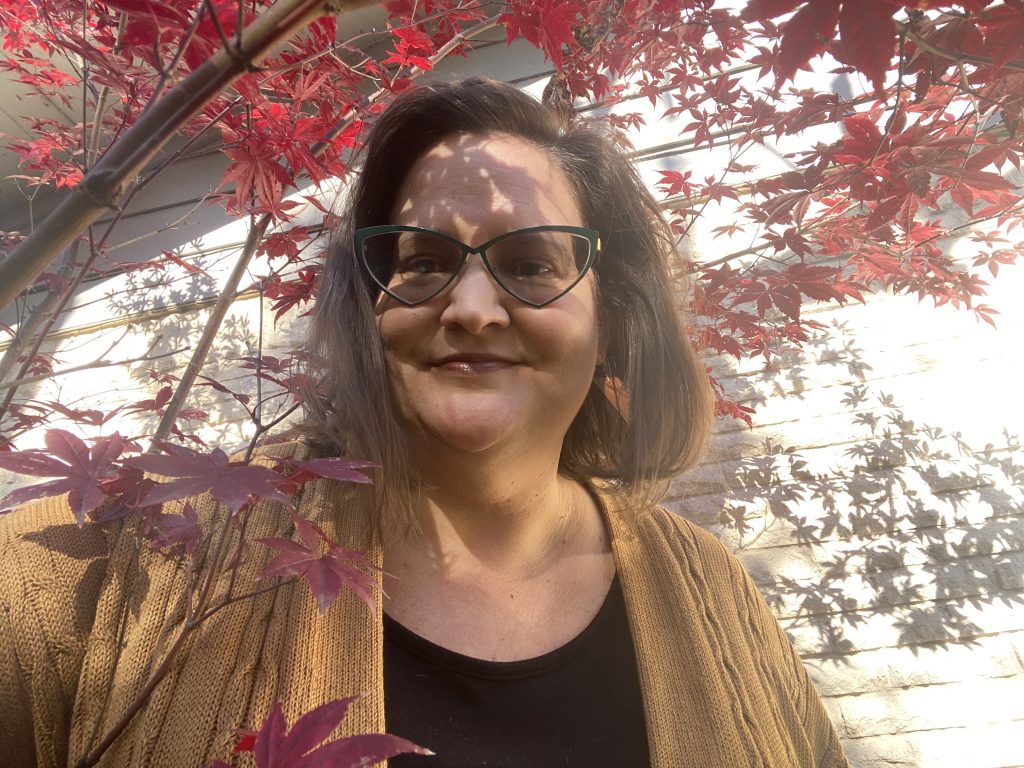 This photo shows a woman with shoulder length brown hair wearing cat-eye style glasses posed smiling under a tree that has red fall leaves.