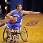 A person wearing a blue basketball jersey holds a basketball while sitting in a wheelchair on a basketball court.