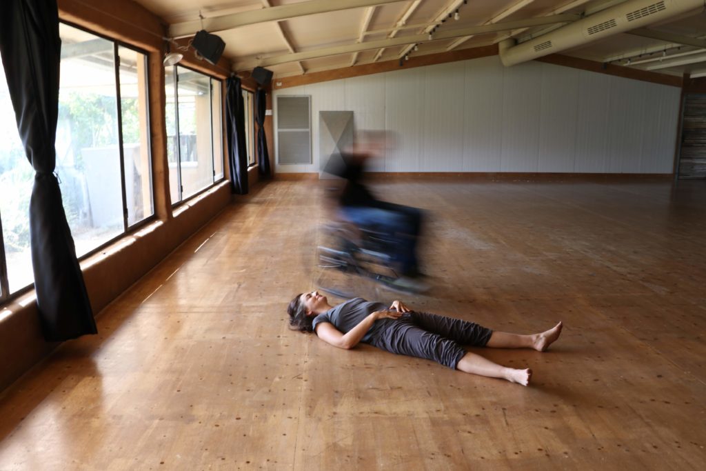 Tali lying on the wood floor with her eyes closed and blurry image of Hai in his wheelchair moving next to Tali.