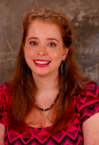 A caucasian young woman with shoulder-length red hair and pink lipstick smiling. She is wearing a pink short sleeved dress with gray triangles and a black zig zag pattern, a black beaded necklace, and hoop earrings. The background is light brown and yellow.