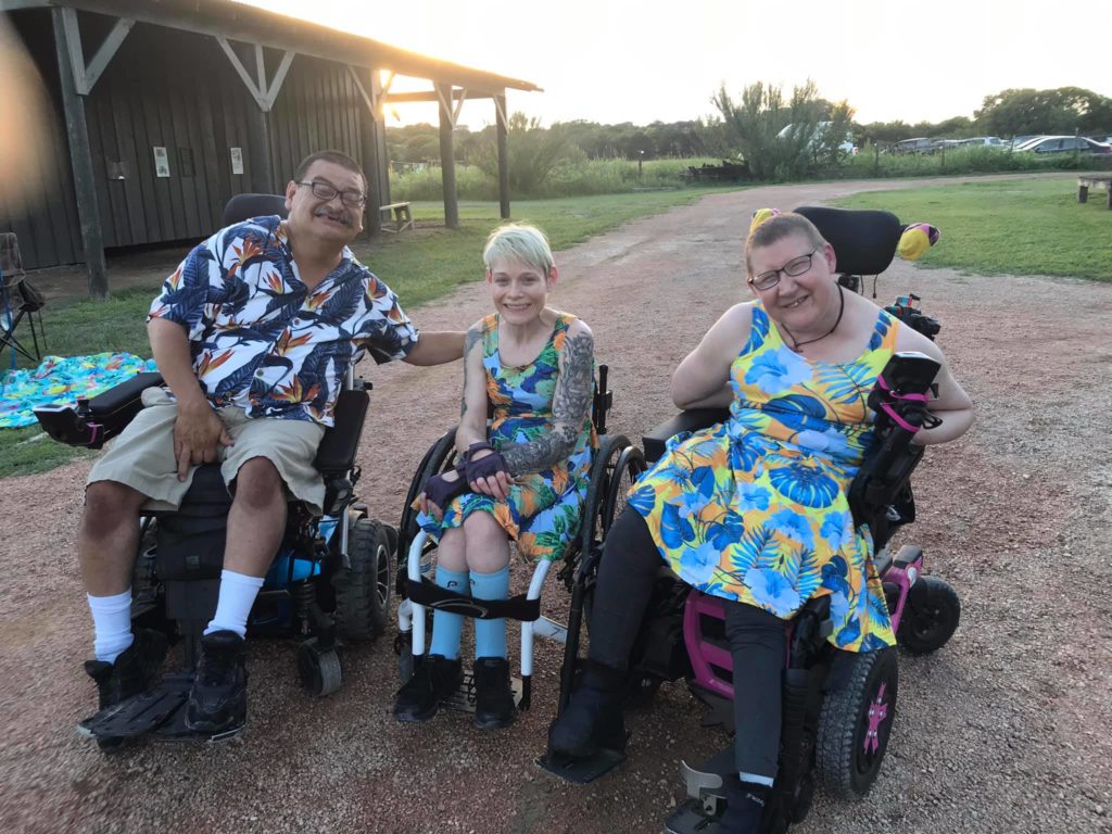 Three people dressed in colorful clothing sit in wheelchairs outside.