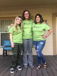 three women in green shirts with the Art Spark Texas logo on the chest pose together with smiles on their faces 